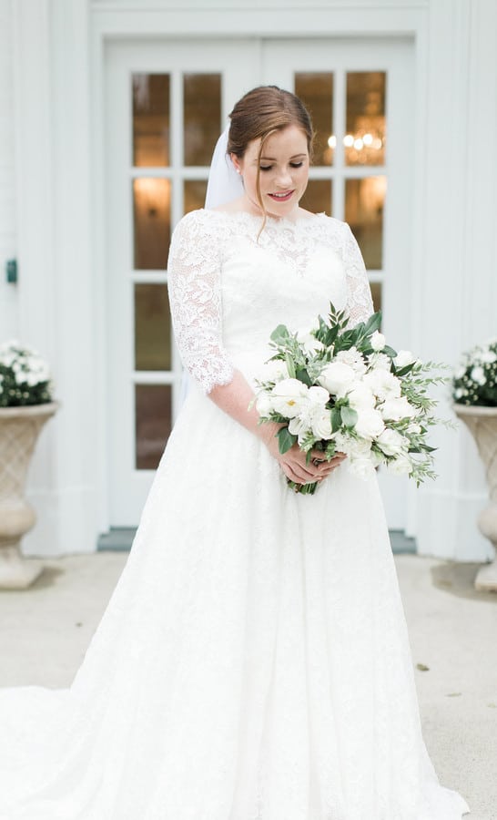 Bride in three-quarter sleeve lace ballgown holding loose white rose bouquet in front of elegant white doors with potted white flowers