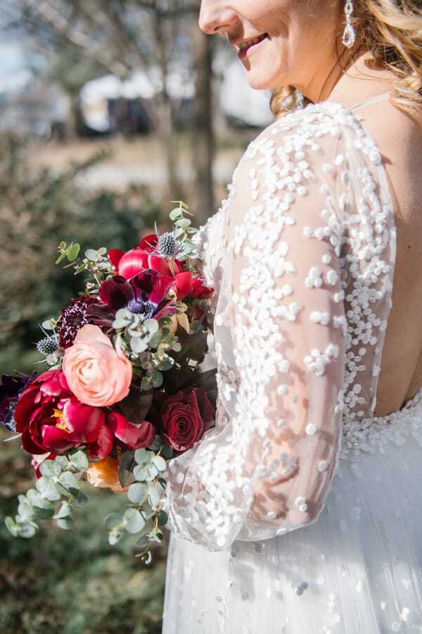 Bride in sheer confetti-dotted sleeve gown holding vibrant bouquet with coral peonies, burgundy anemones, pink ranunculus, and blue thistle