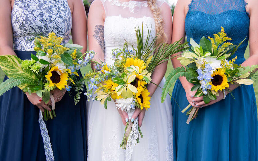 Bride and bridesmaids in navy and teal dresses holding wildflower sunflower bouquets with blue delphinium, solidago, and ferns