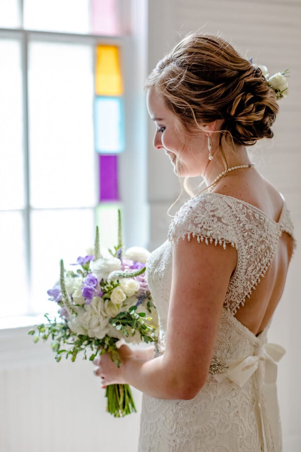 Profile view of bride smiling at bouquet of white roses, purple freesia, and lavender sprigs near colorful stained glass