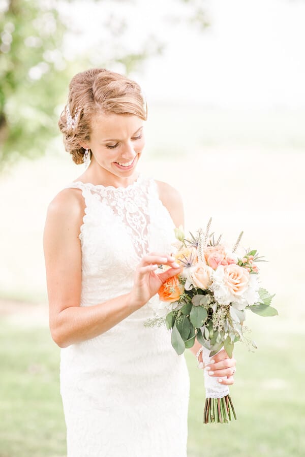 Bride in high-neck lace gown looking at her wedding ring while holding peach and coral rose bouquet with blue thistle accents