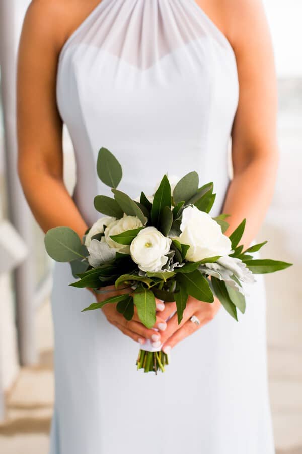 Bridesmaid in pale blue halter dress holding petite bouquet of white ranunculus, roses, dusty miller, and mixed greenery