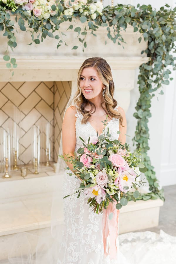 Bride in v-neck lace gown smiling in front of eucalyptus-draped fireplace holding large soft pink bouquet with roses, orchids, and trailing blush ribbons