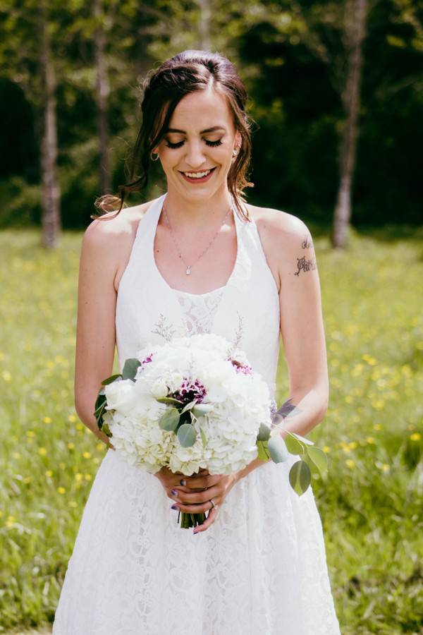 Smiling bride in halter top A-line gown holding white hydrangea bouquet with purple accents in sunny field