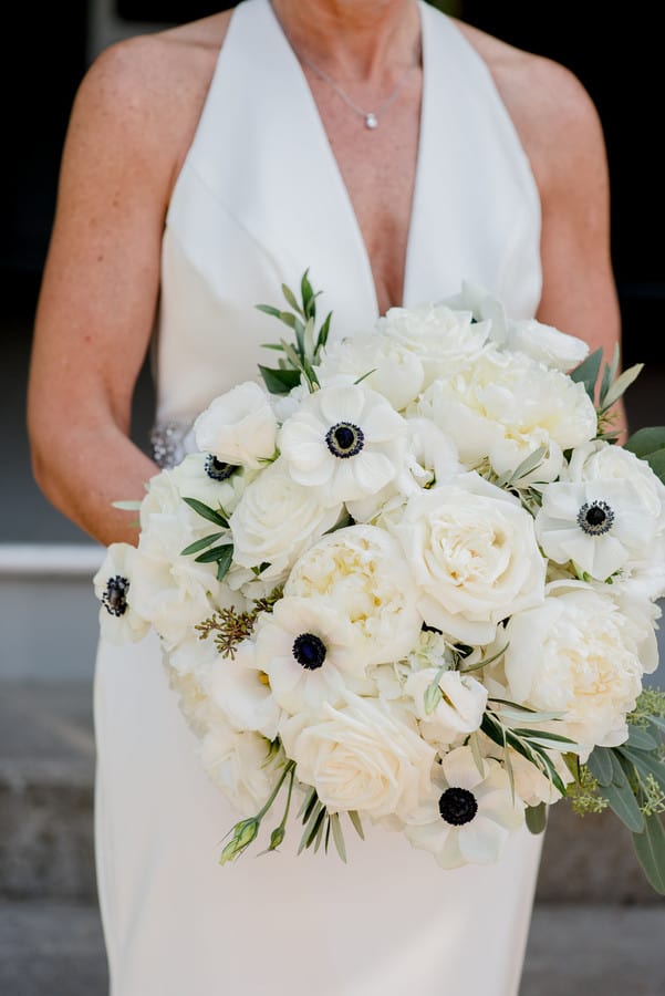 Bride in halter satin gown holding compact round bouquet of white roses, peonies, and anemones with black centers and olive branch greenery