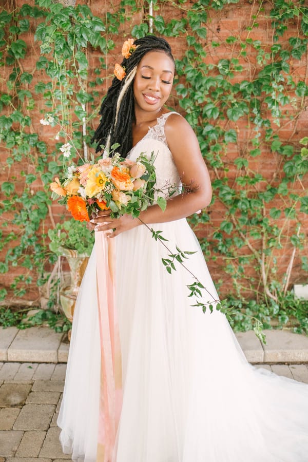 Joyful bride in tulle a-line gown with locs adorned with orange flowers holding sunset-toned bouquet of orange ranunculus, peach roses, and yellow blooms against ivy-covered brick wall