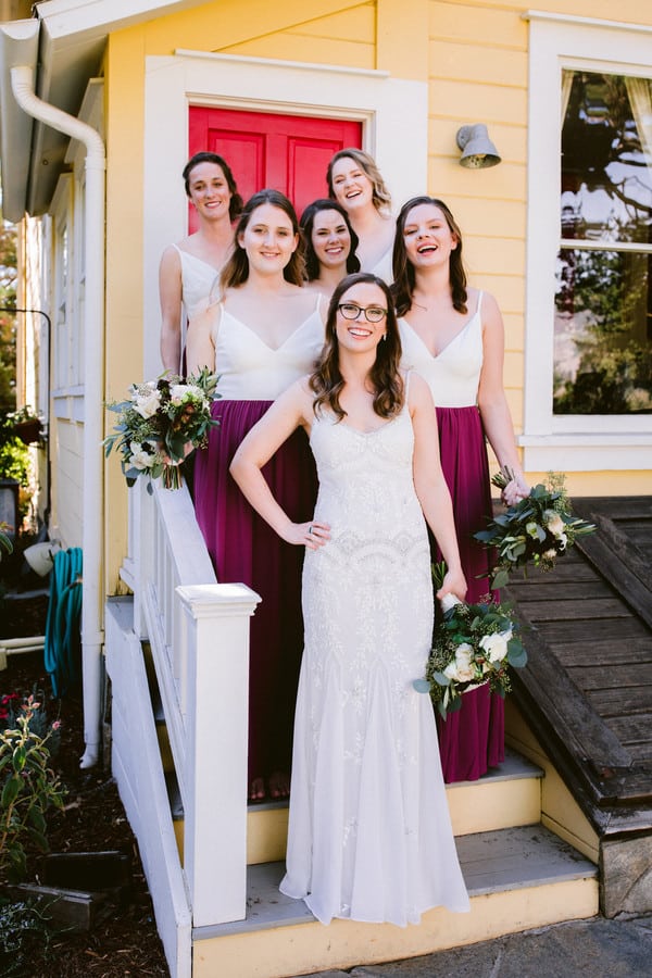 Bride on yellow farmhouse porch steps with bridesmaids wearing two-tone cream and burgundy dresses holding white and green bouquets