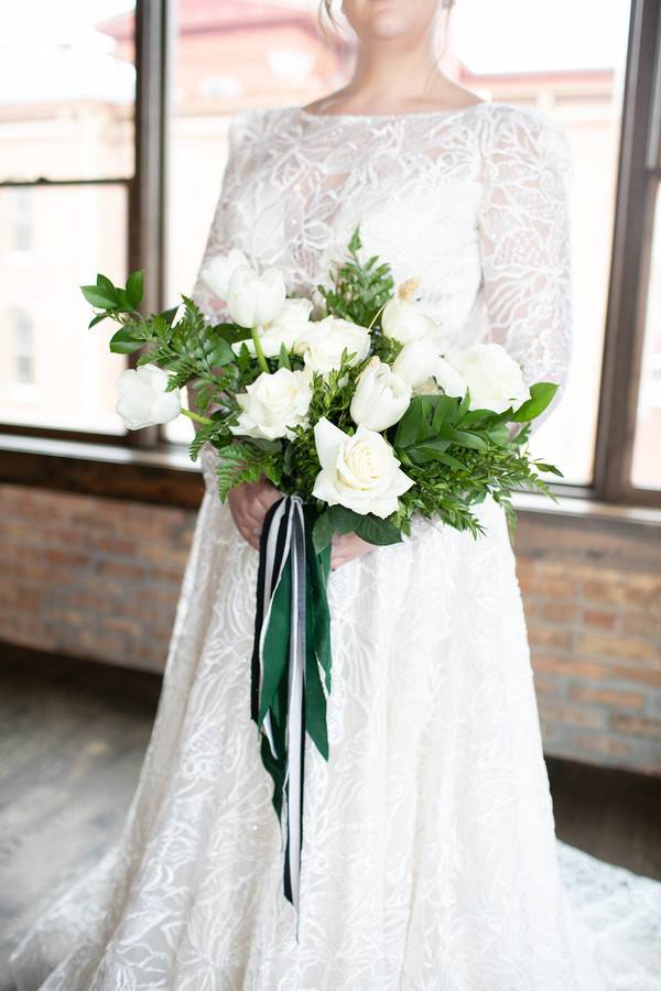 Bride in long-sleeve lace ballgown holding white rose and tulip bouquet with emerald green and white ribbon streamers against industrial brick window