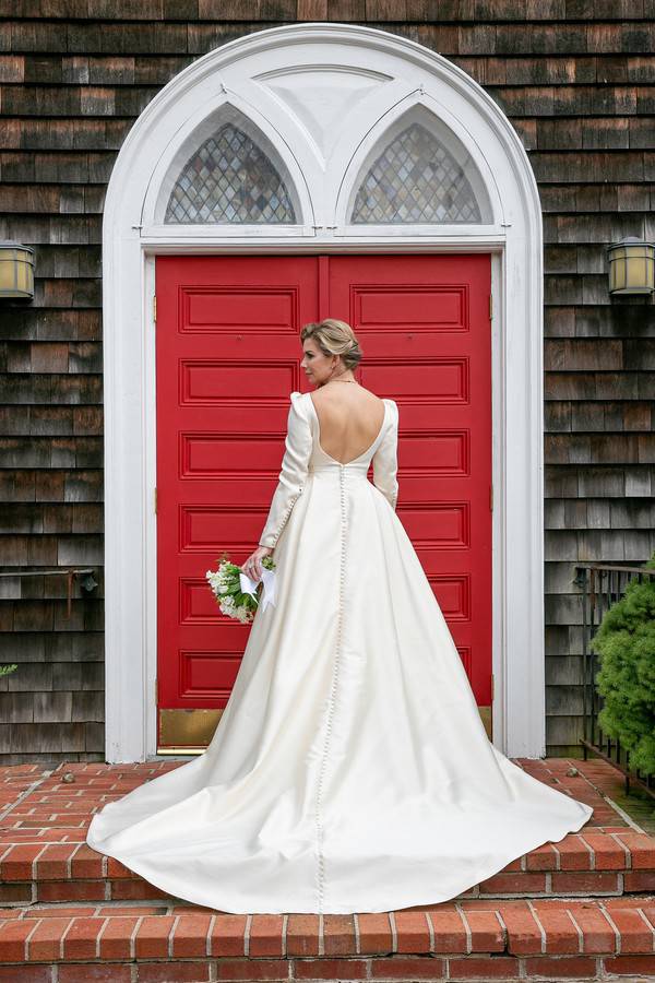 Bride facing away showing deep V-back of long-sleeve satin ballgown with covered buttons against red church door