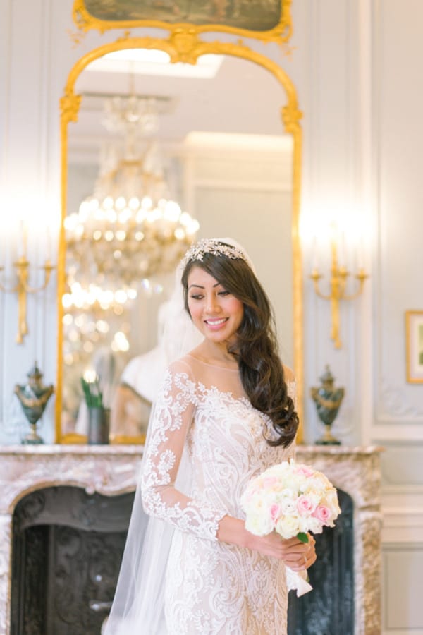 Bride in illusion lace gown with crystal headpiece holding compact pink and white rose bouquet in luxurious Parisian room with gold mirror and chandelier