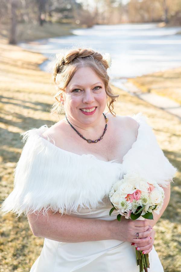 Bride in white satin off-shoulder A-line gown with fluffy white fur stole holding blush and white bouquet with snowy Colorado landscape behind