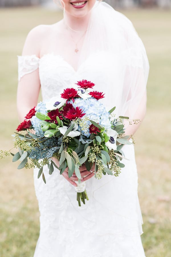 Bride in off-shoulder lace gown holding loose bouquet with burgundy chrysanthemums, pale blue hydrangeas, white anemones, and seeded eucalyptus