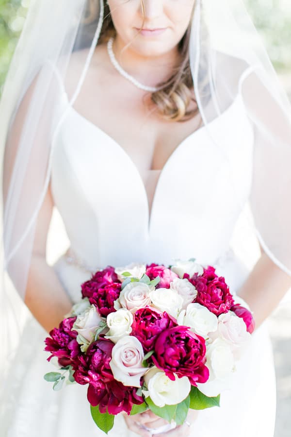 Bride in plunging V-neck gown with veil holding compact bouquet of deep magenta peonies and white roses with touches of greenery