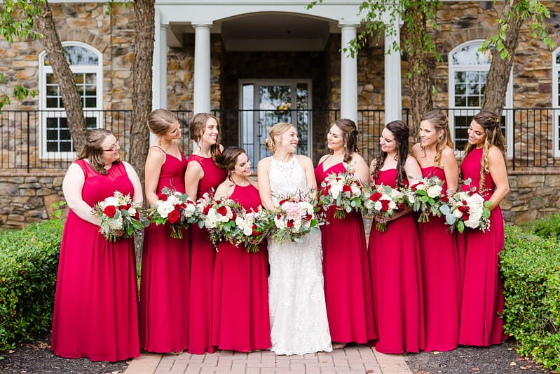 Bride surrounded by eight bridesmaids in various styles of red and burgundy dresses holding matching bouquets of red roses and white hydrangeas at stone manor venue