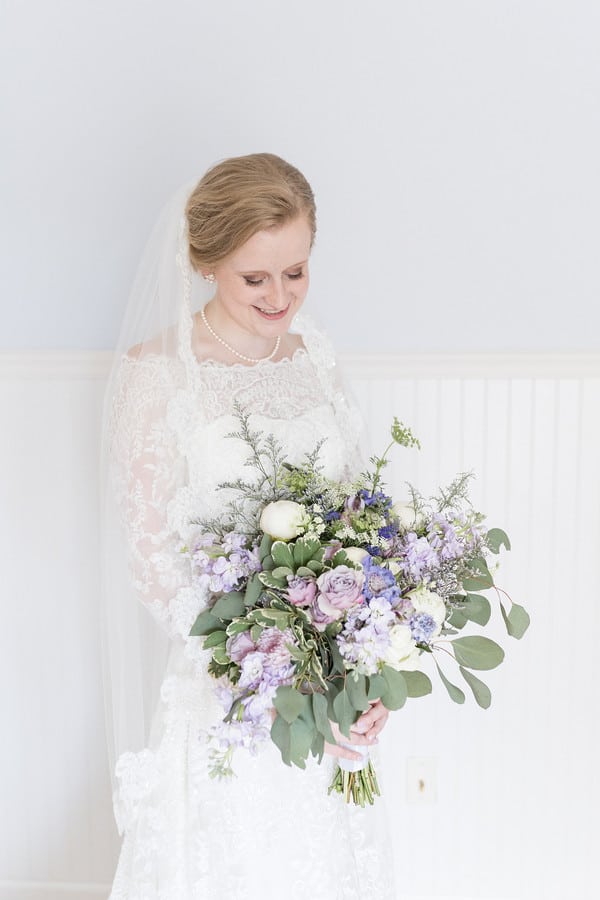 Bride in long sleeve illusion lace gown looking down at lush bouquet of lavender roses, blue delphinium, eucalyptus, and purple stock