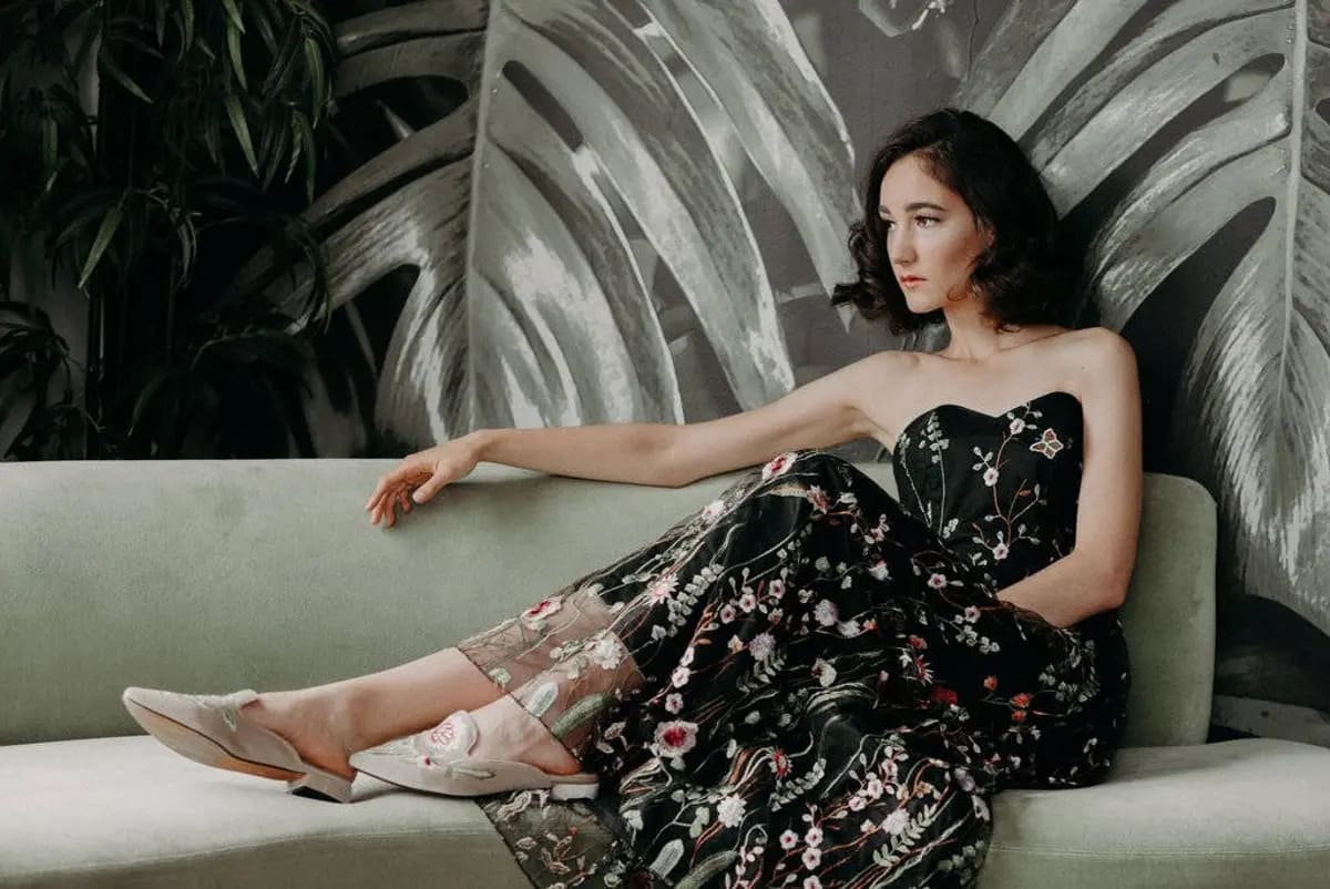 A woman seated on a light-colored couch against a backdrop of large, grayscale palm leaves. She wears a strapless dress adorned with colorful floral patterns. The woman has short, wavy hair and is gazing off to the side. Her feet are clad in white shoes with floral embellishments. The overall ambiance of the picture is serene and elegant, with a touch of vintage charm.