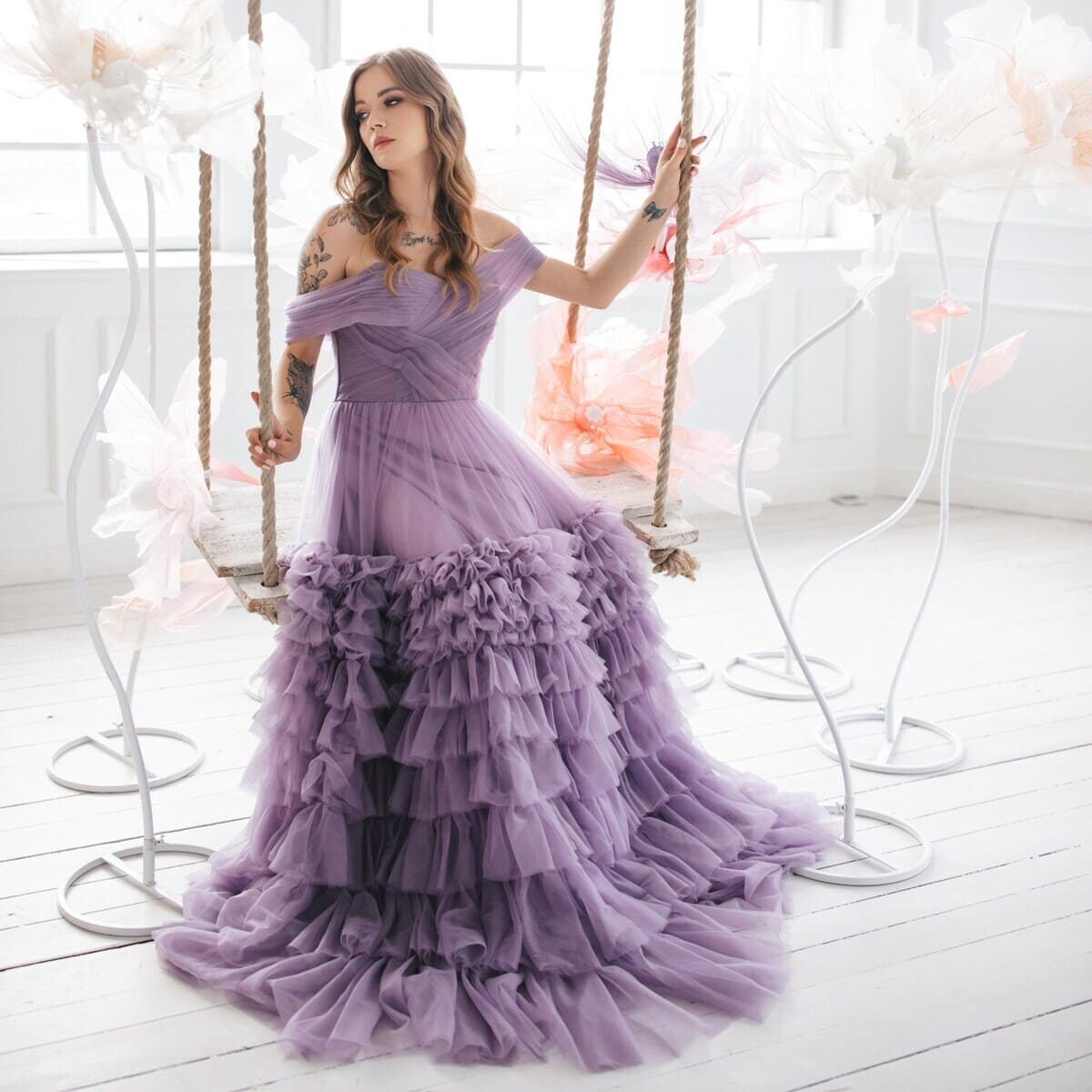 A woman seated on a swing, surrounded by oversized, translucent flowers. She wears a voluminous, lavender-colored gown with ruffled layers. The setting is a bright, airy room with large windows, white walls, and a wooden floor. The swing she's on has thick ropes, and the flowers are positioned both around her and in the background.