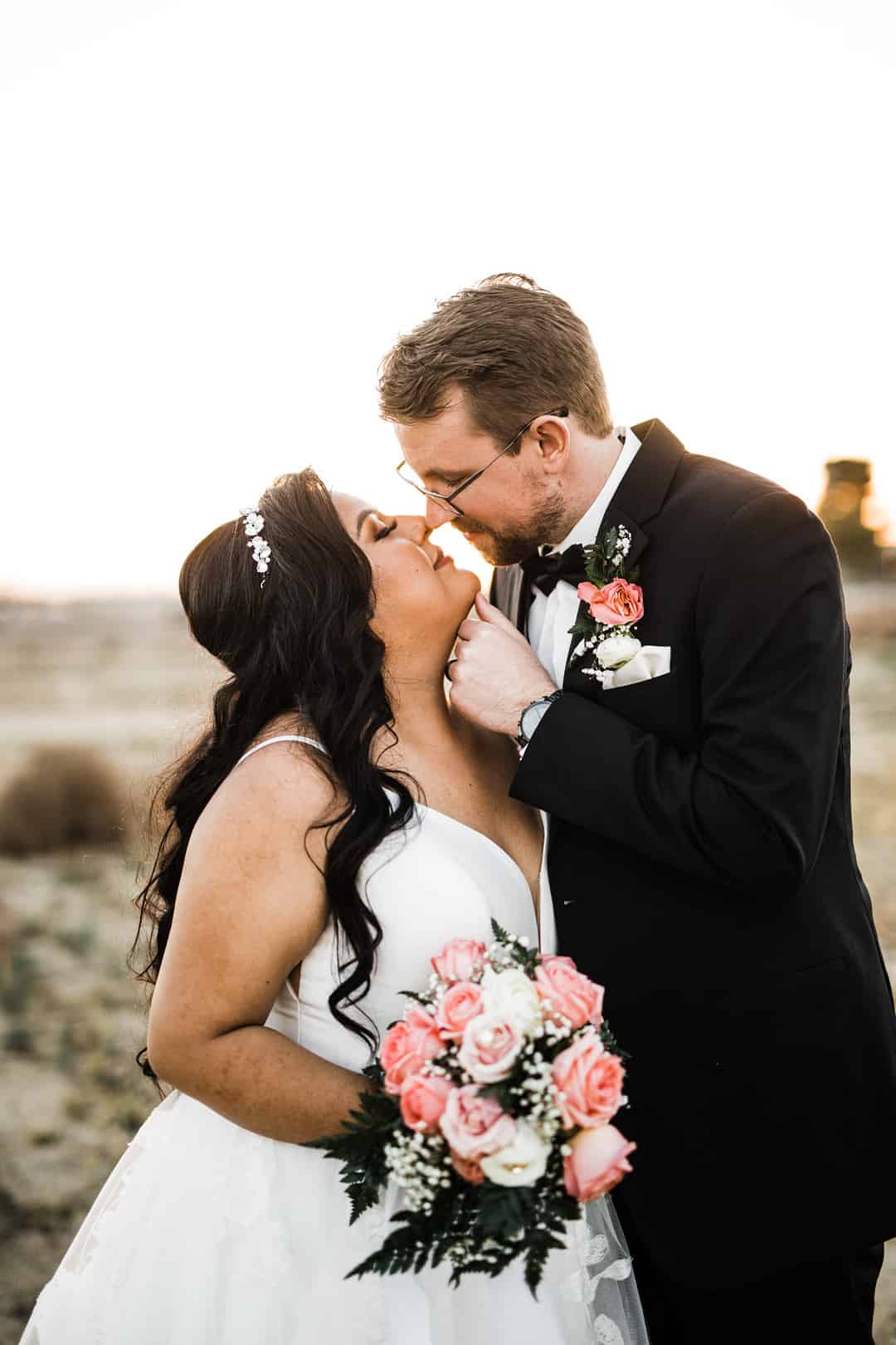 Groom in black suit lifting bride's chin for kiss at sunset, bride holding classic pink and white rose bouquet with baby's breath and ferns