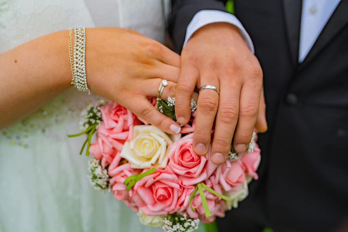 Close-up of a bride and groom's hands with wedding rings resting on a bouquet of pink and white roses, highlighting their thoughtful flower choices for meaningful wedding flowers.