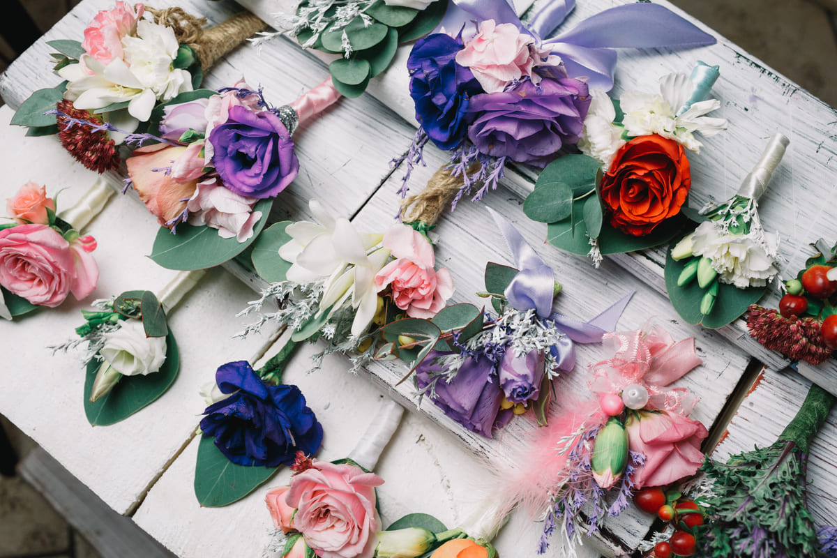 A variety of colorful boutonnieres with roses, lilies, and greenery—perfect wedding flowers—are arranged on a white wooden surface.