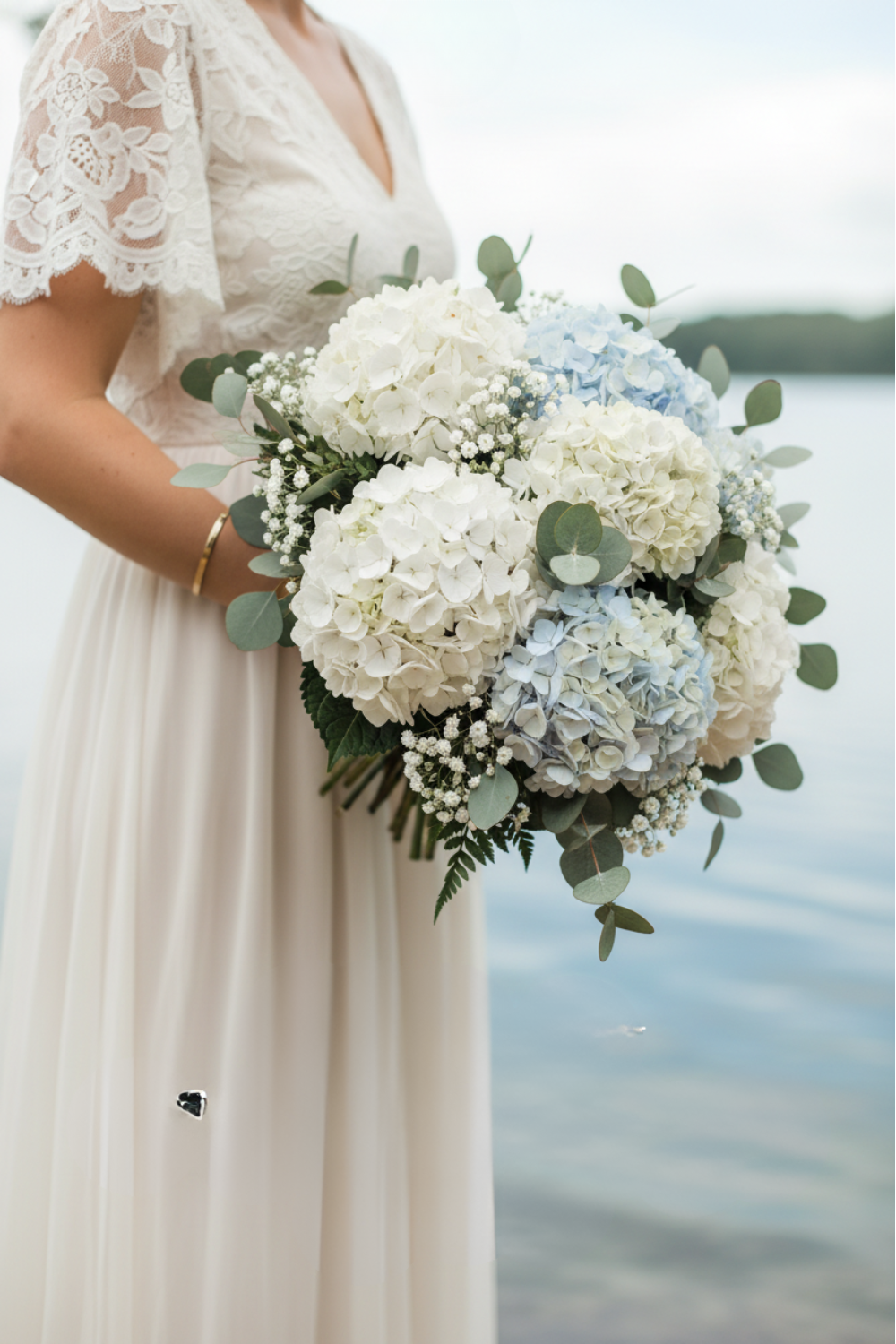 Bride in lace-sleeve gown holding white and blue hydrangea bouquet with eucalyptus and baby's breath by the water