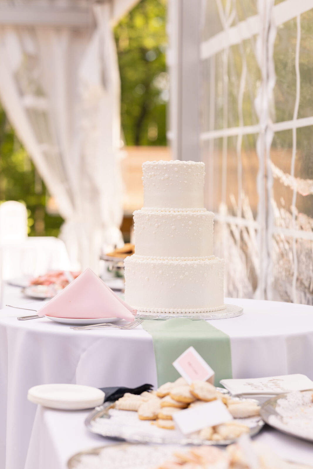 A three-tiered white wedding cake with pearl-like decorations sits on a table with cookies, a pink napkin, and pastel-colored place cards in an outdoor setting.