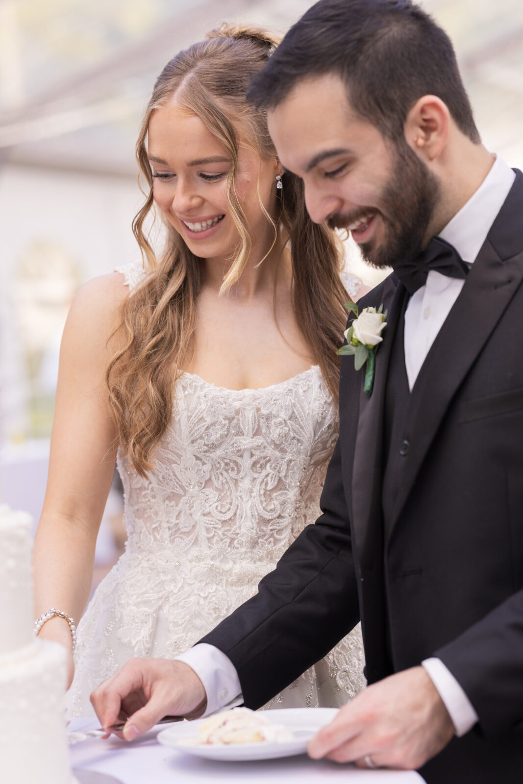 A bride and groom, dressed in formal wedding attire, smile as they cut a wedding cake together at their reception.
