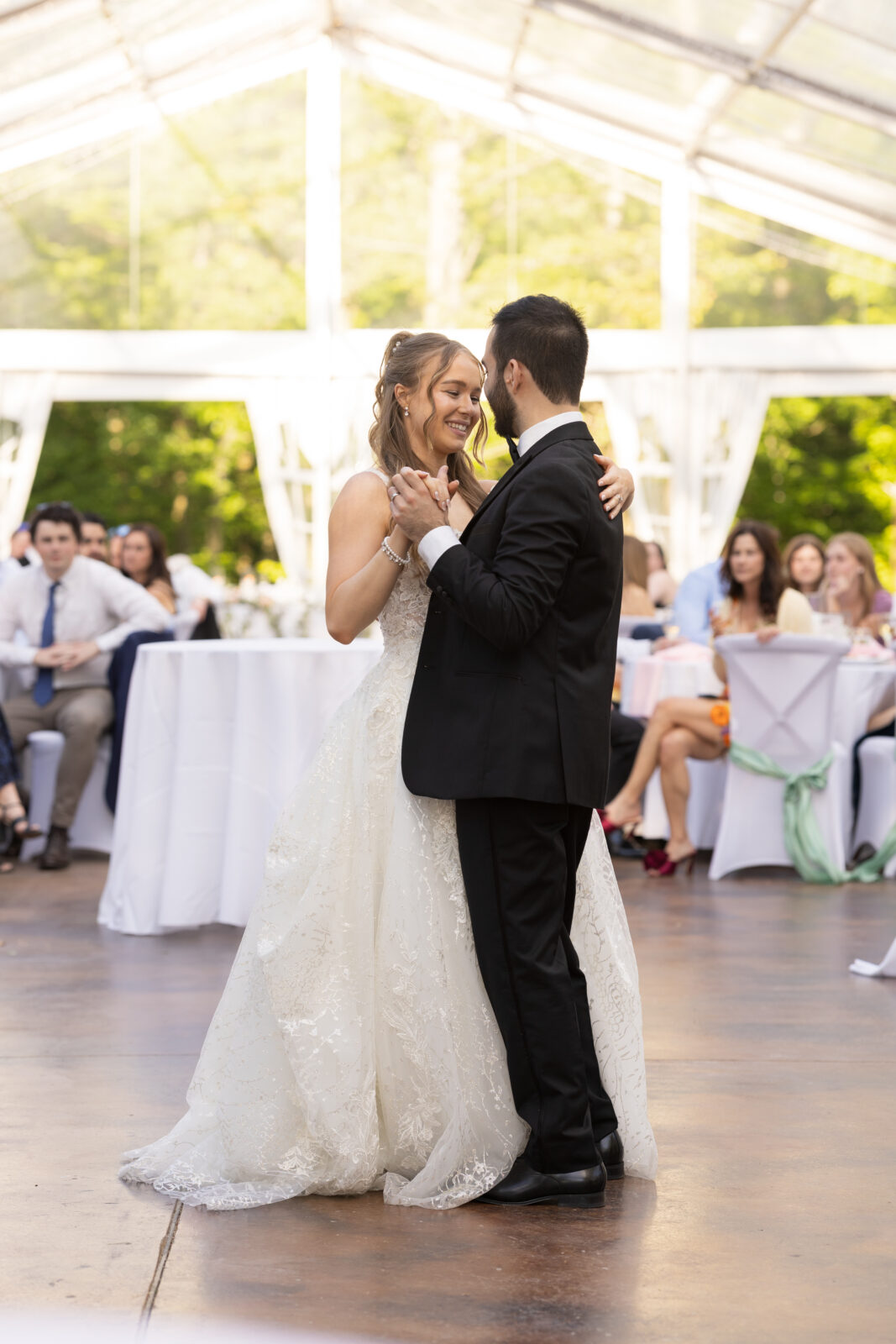 A bride and groom share their first dance at a wedding reception, surrounded by seated guests under a clear tent.
