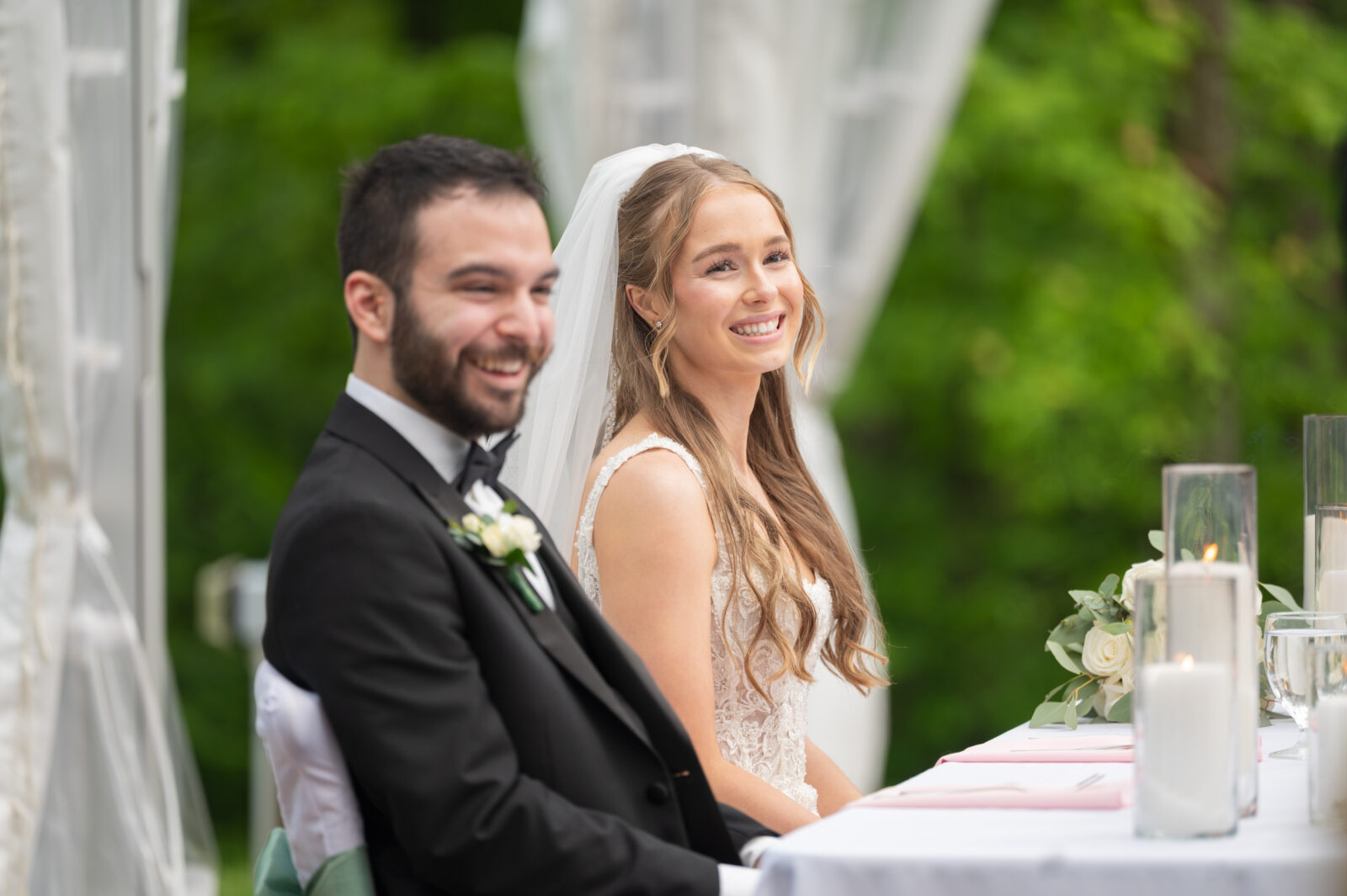 A bride and groom sit side by side at a table, smiling, dressed in wedding attire with candles and greenery in the background.