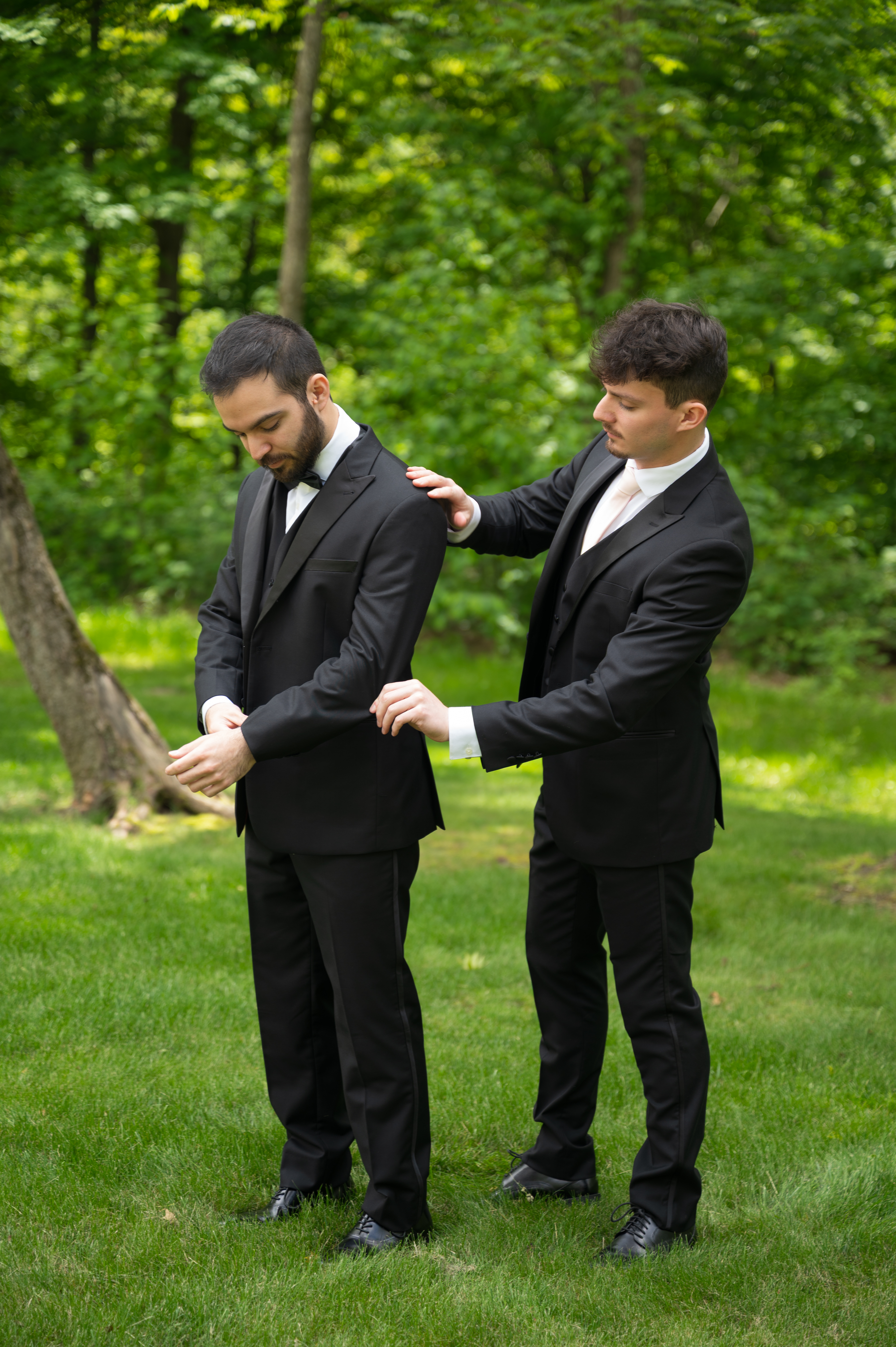 Groom in black tuxedo adjusting his cuff while a groomsman in a black tuxedo steadies his shoulder on a green lawn