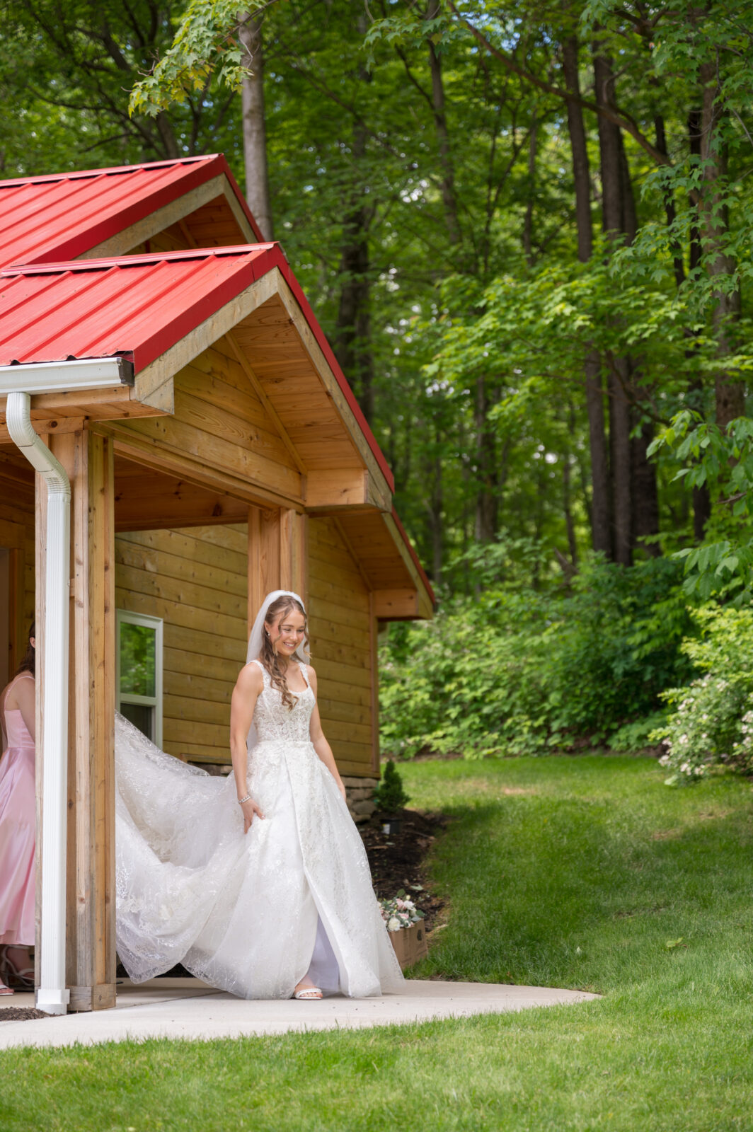 A bride in a white dress and veil steps out of a wooden building with a red roof, surrounded by greenery on a sunny day.