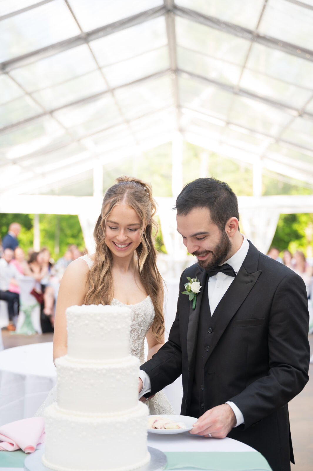 A bride and groom in formal attire stand together cutting a white tiered wedding cake at a reception under a clear tent.