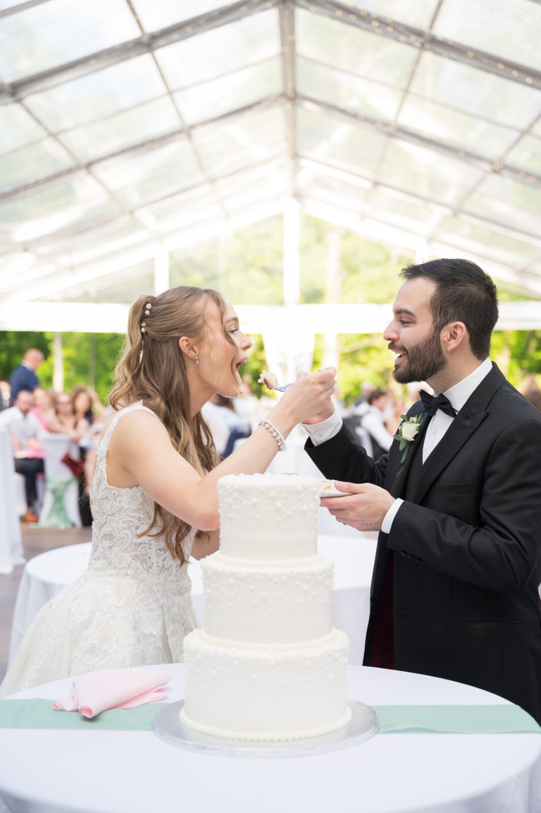 A bride and groom feed each other cake at their wedding reception, standing by a three-tiered white cake under a clear tent.