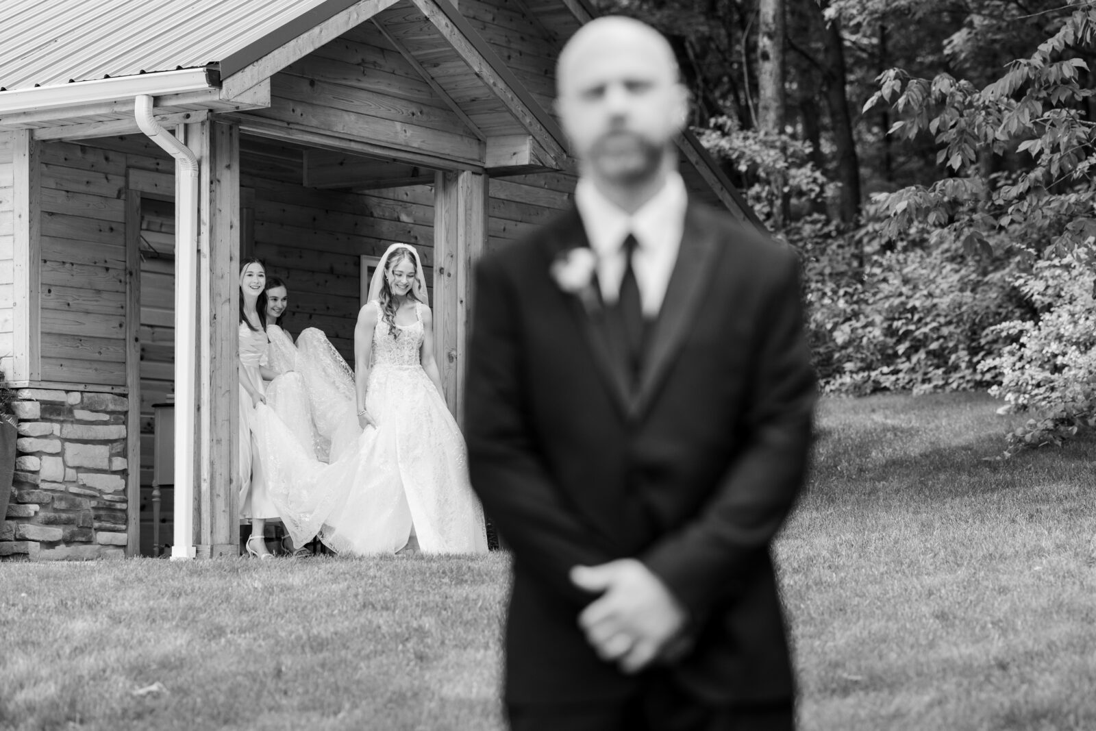 A bride in a wedding dress, accompanied by two women, approaches a suited man standing outside a wooden building; the man is out of focus in the foreground.