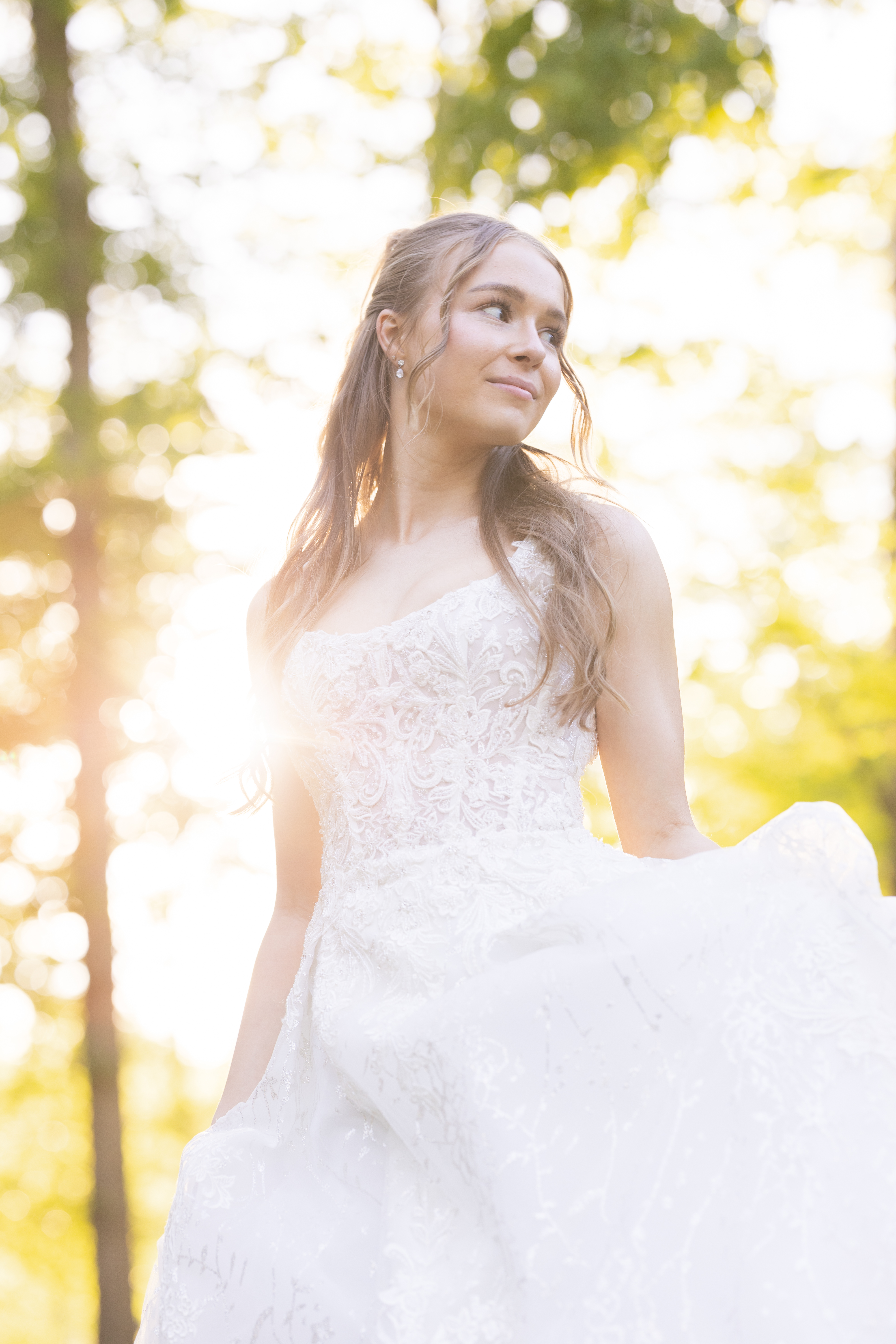 Bride in a one-shoulder lace ball gown backlit by golden sunlight in the forest, gathering her skirt and looking off to the side