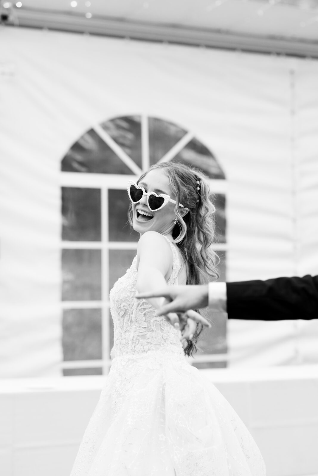 A bride wearing heart-shaped sunglasses smiles and looks back while holding hands with someone, possibly dancing, in a white tented venue.