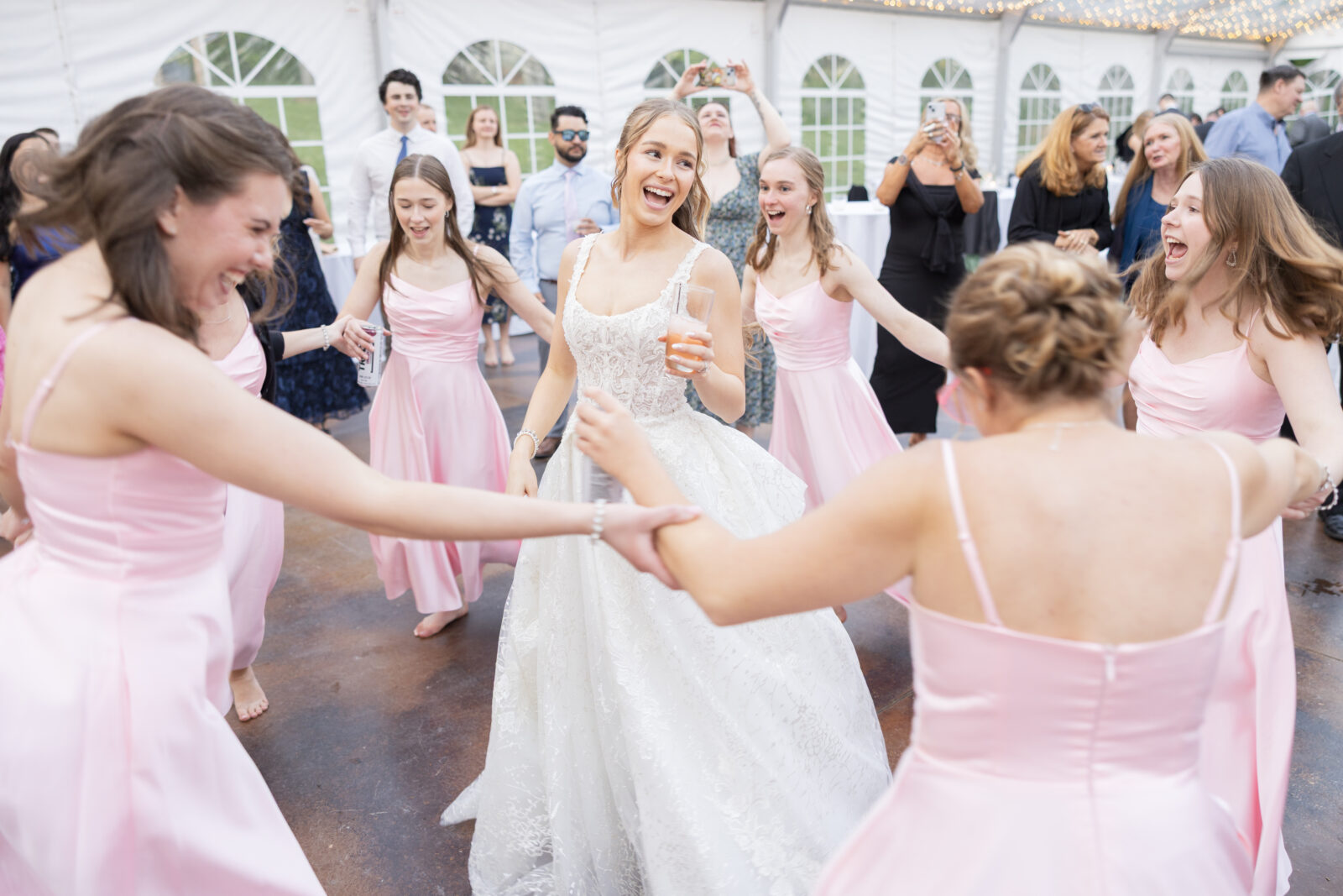 A bride in a white dress dances in a circle with bridesmaids in pink dresses at a wedding reception under a white tent.