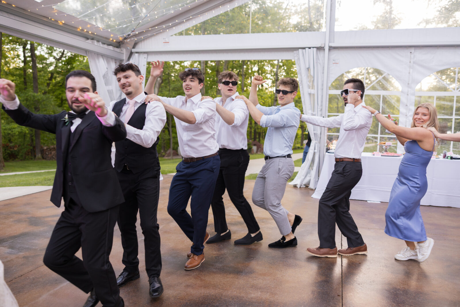 A group of people, mostly men in dress shirts and trousers, and one woman in a blue dress, dance in a conga line at an indoor-outdoor event space.