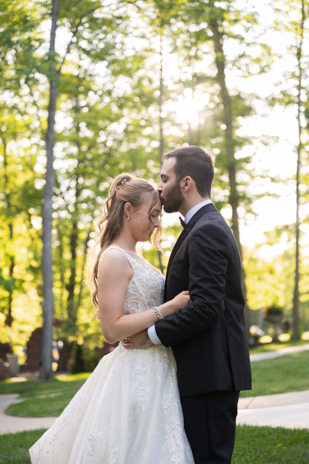 A bride and groom stand close outdoors, with the groom kissing the bride’s forehead. Sunlight shines through the trees in the background.