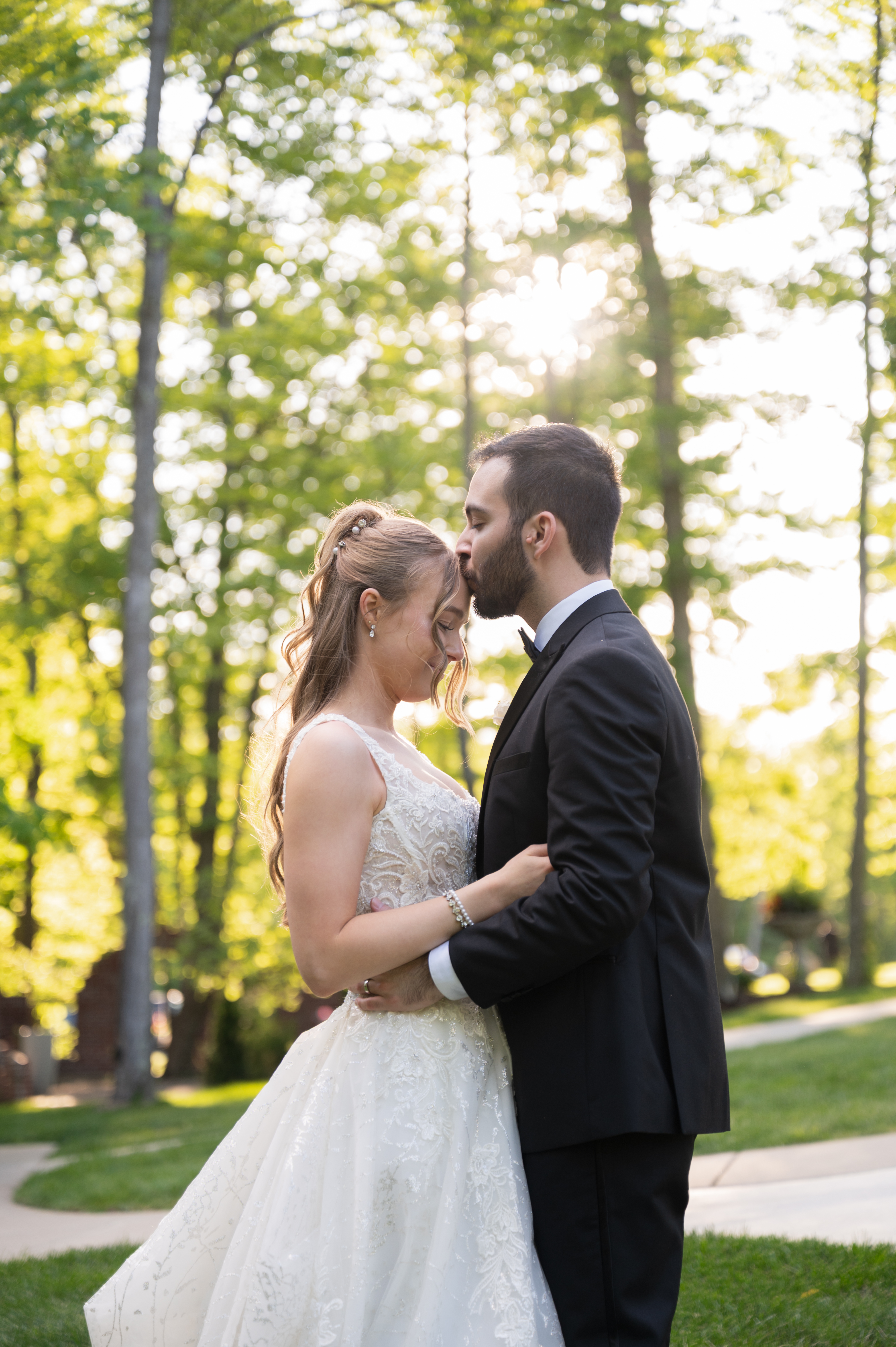 Groom kissing the bride's forehead outdoors in a sunlit wooded setting, both eyes closed
