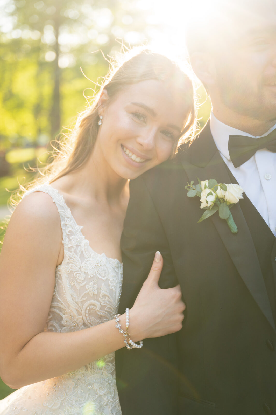 A bride in a lace dress smiles and leans on a groom in a black tuxedo, with sunlight shining behind them in an outdoor setting.
