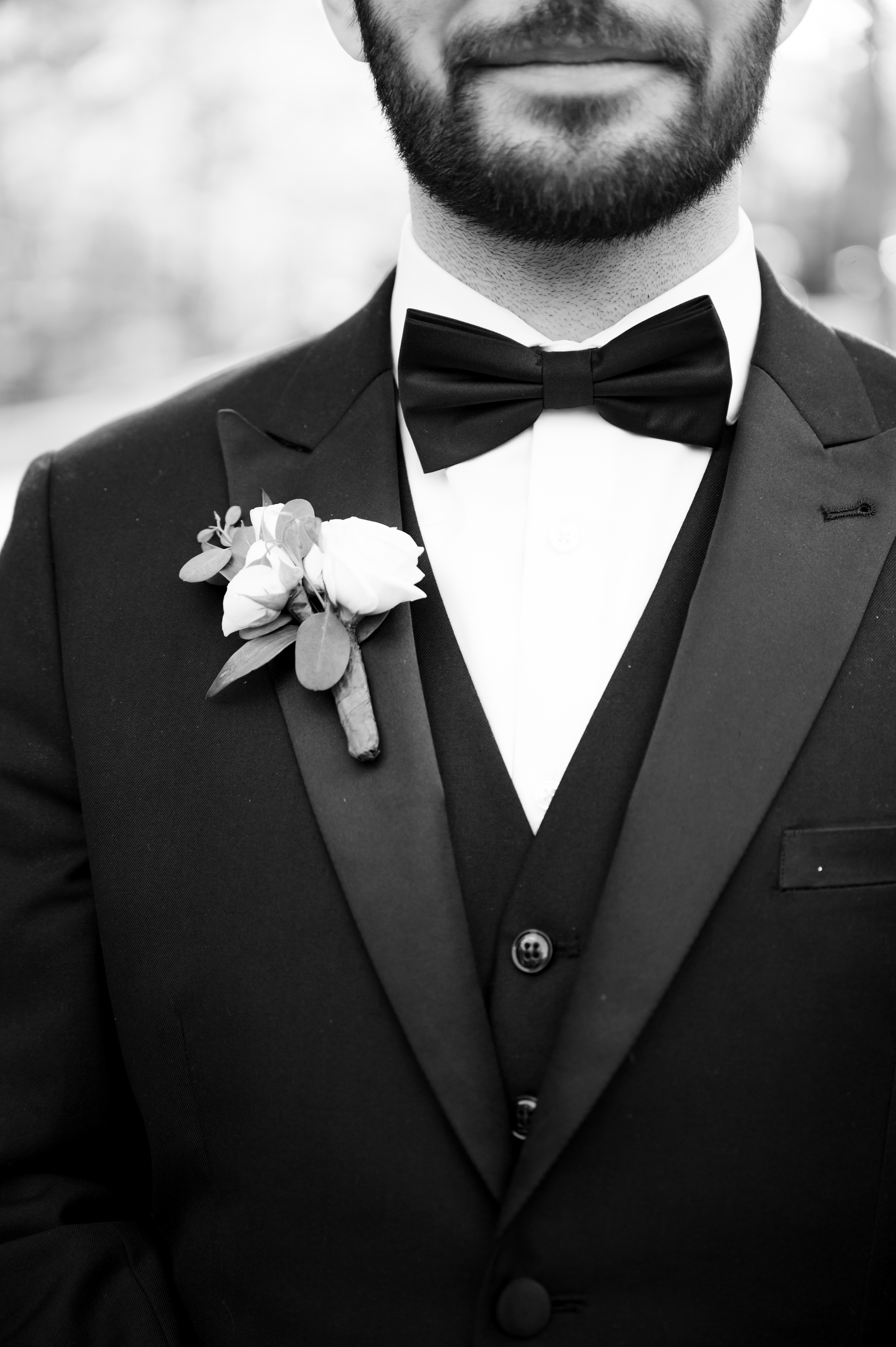 Black and white close-up of a groom in a black three-piece tuxedo with satin peak lapel, black bow tie, and white rose boutonniere