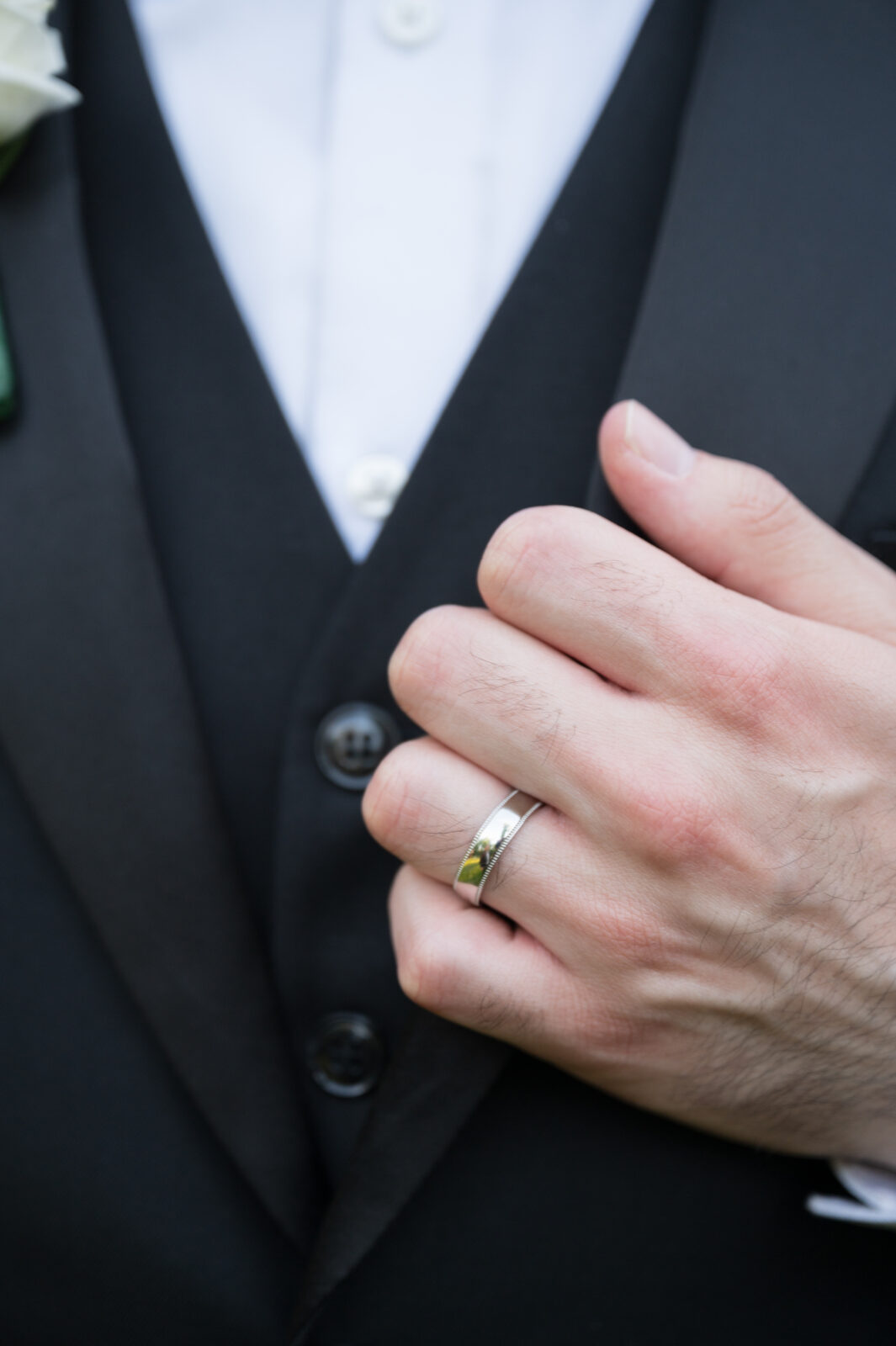 Close-up of a person in a black suit and white shirt, wearing a silver ring on their left ring finger and adjusting their jacket.