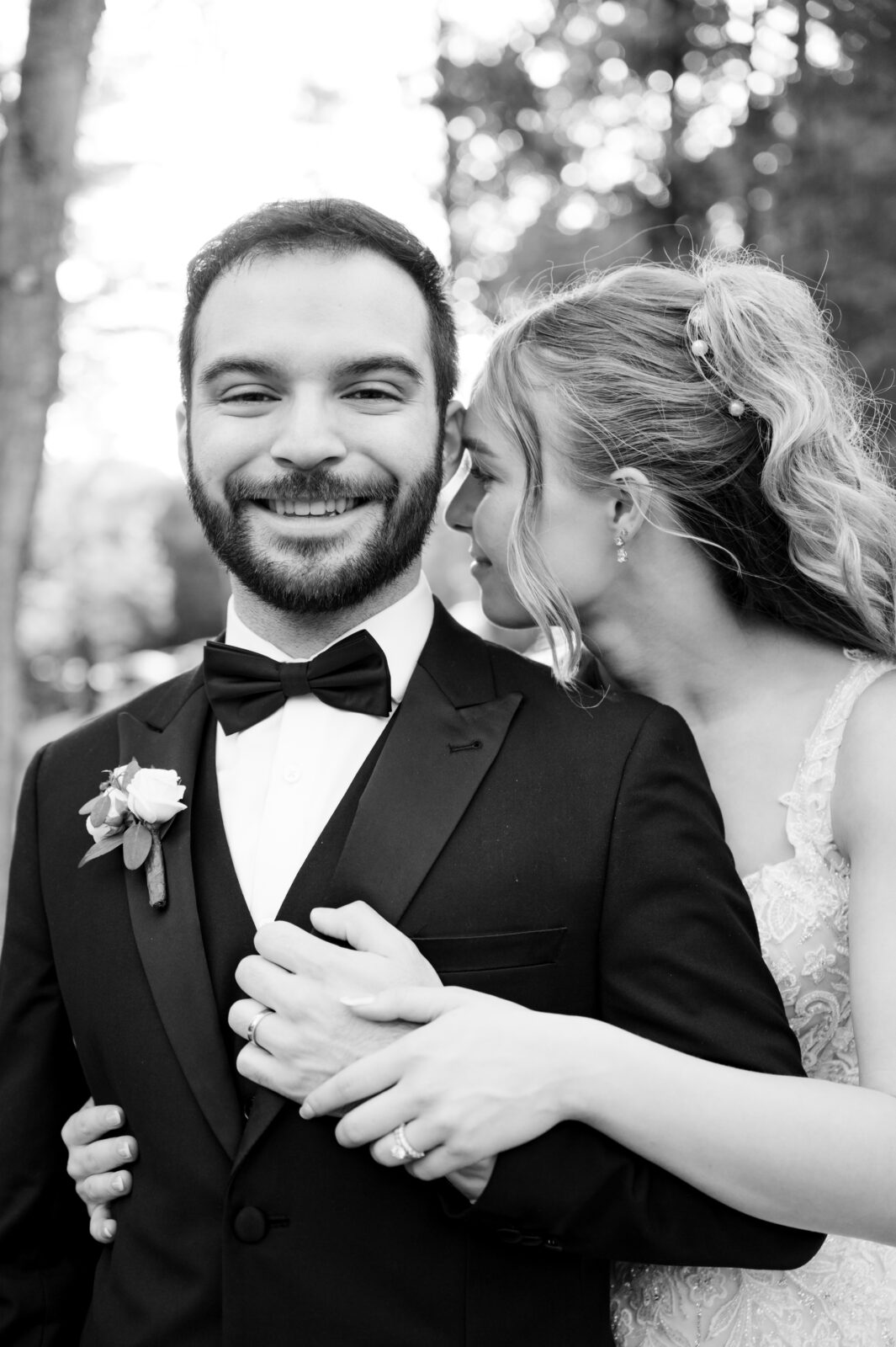 A bride embraces a groom from behind, both smiling, in an outdoor setting. The groom wears a tuxedo with boutonniere; the bride wears a lace dress. The image is in black and white.