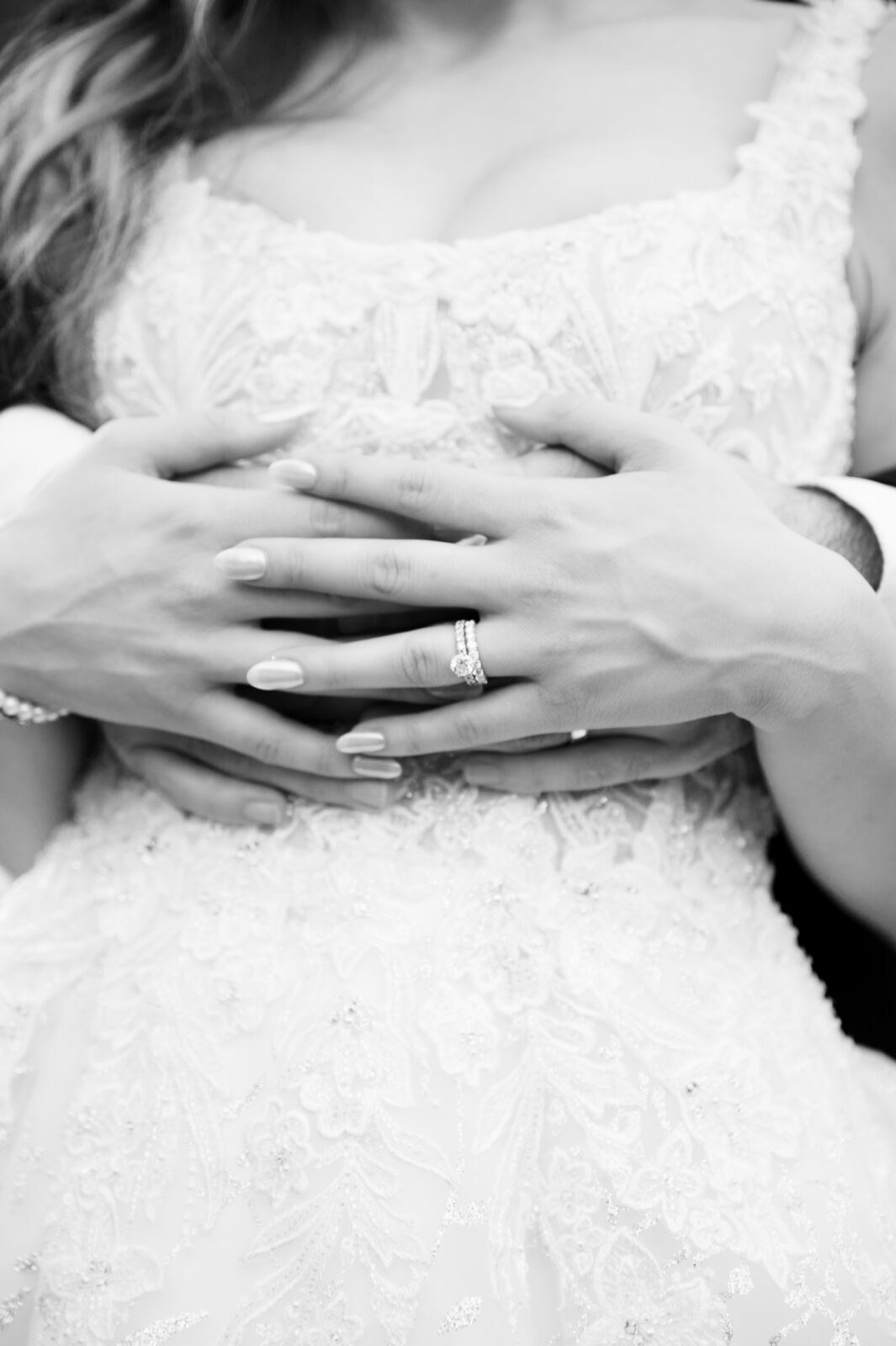 Close-up of a couple's hands in an embrace, with a wedding ring visible on the woman's finger, over a detailed lace wedding dress.