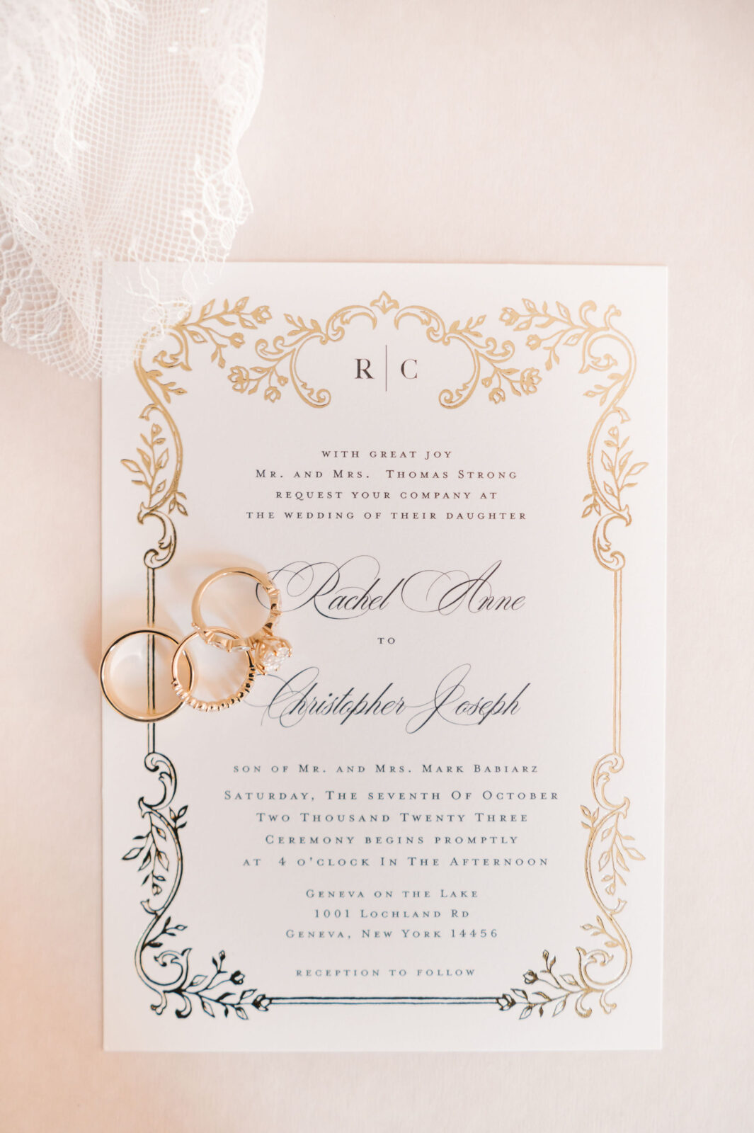 A wedding invitation card with gold decorative borders and two gold rings placed on it; a piece of white lace fabric is in the top left corner.
