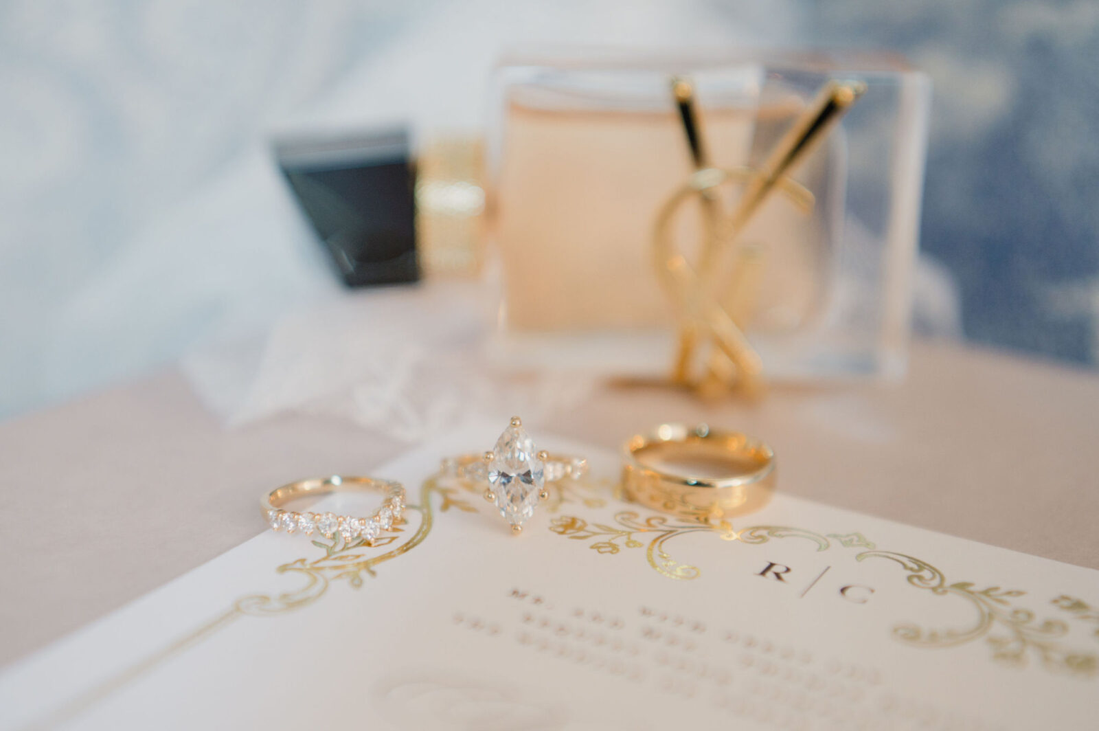Three gold rings and a perfume bottle are arranged on a decorated invitation card with initials "R | C" printed at the top.