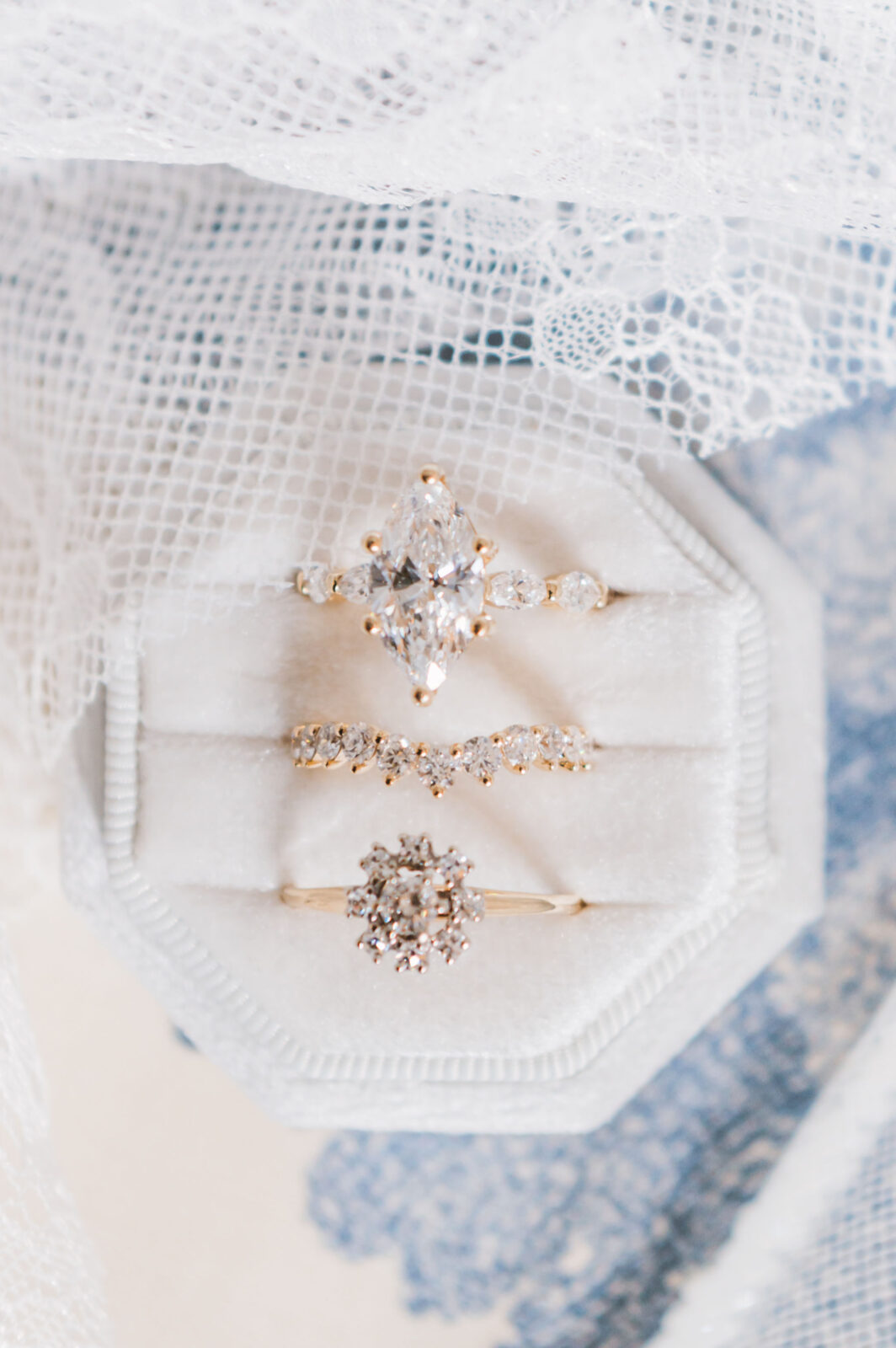 Three gold rings with diamond settings displayed in a white velvet ring box, placed on lace fabric.