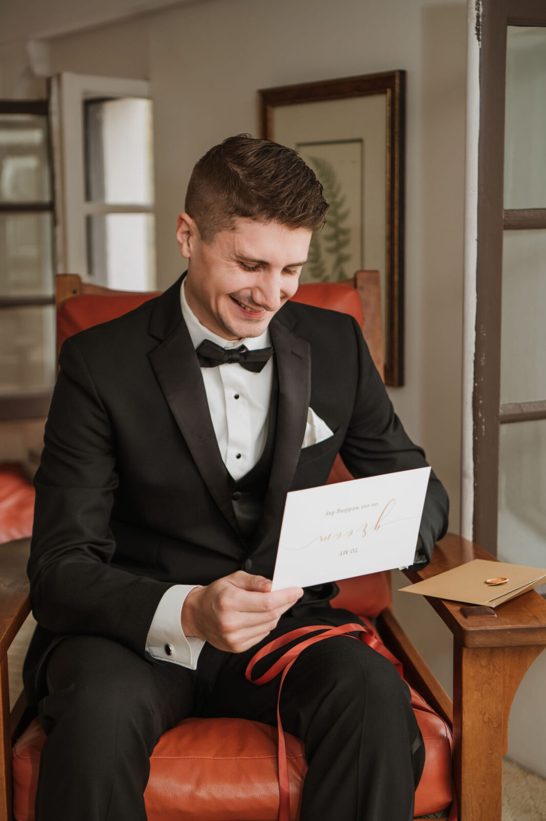 A man in a black tuxedo sits on a chair, smiling while holding and reading a card with an envelope and a ring box on the armrest beside him.