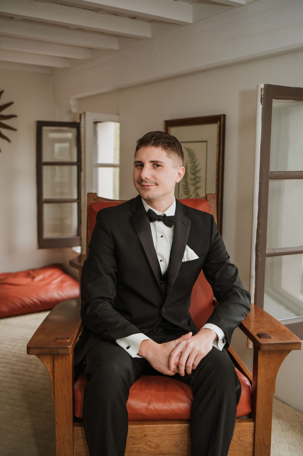 A man in a black tuxedo sits on a wooden chair in a room with open windows and framed artwork on the walls.